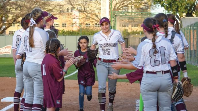 Gina Monaco in her uniform runs alongside a little girl while 澳门金沙线上赌博官网 垒球 players and other little girls form a line on either side of them, offering high fives during a trinity 垒球 game