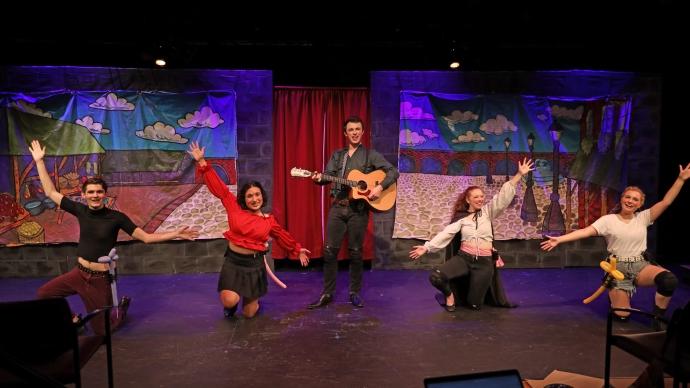 five performers on stage, one in the center holding a guitar, dance to a musical number in the Attic Theater